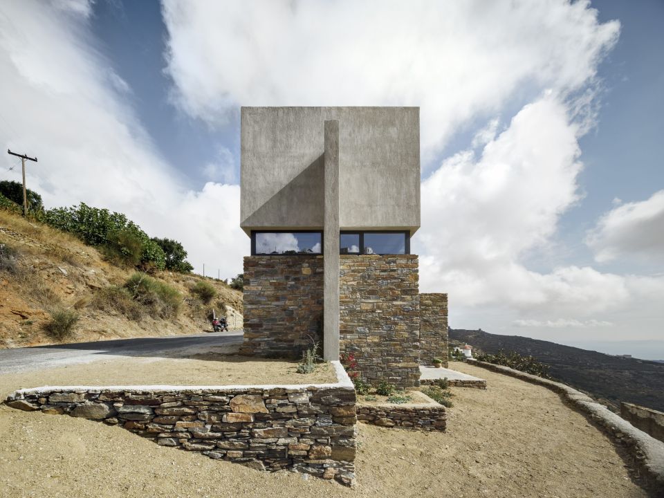 A Touch of New: concrete box on a stone base beside a mountain road, with a thin concrete column and cloudy sky