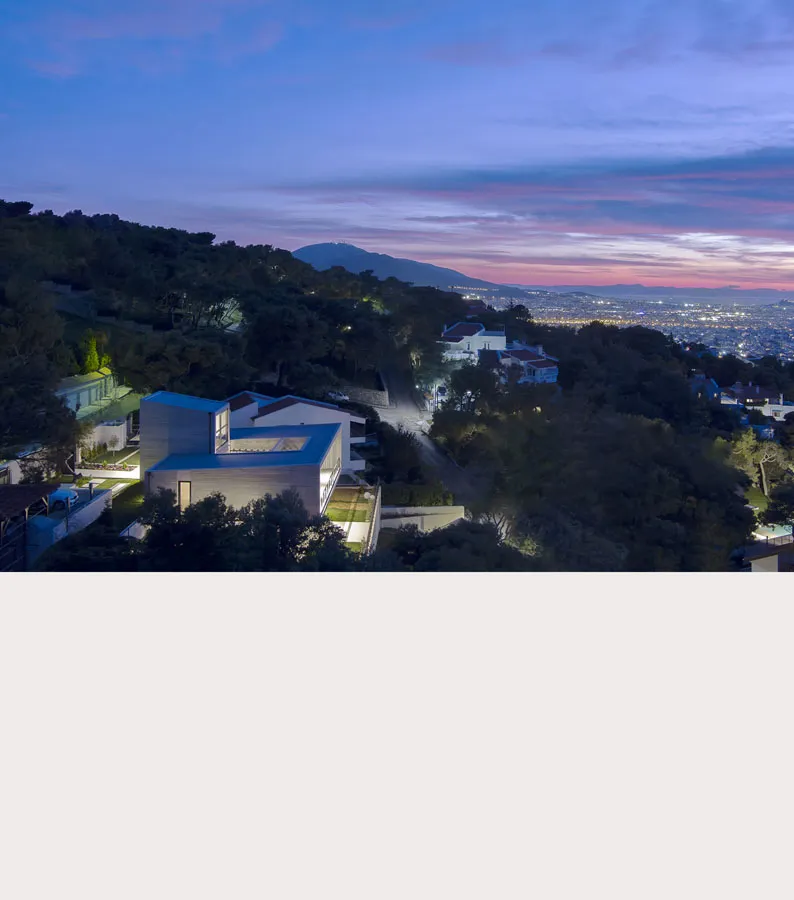 Aerial dusk view of a modern house with a blue roof on a wooded hillside above the lights of Athens