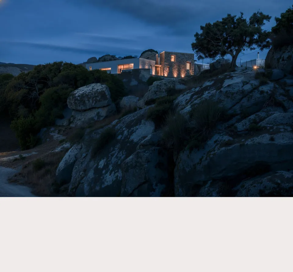Stone and white-rendered house glowing at dusk on a rocky ridge beside a lone tree