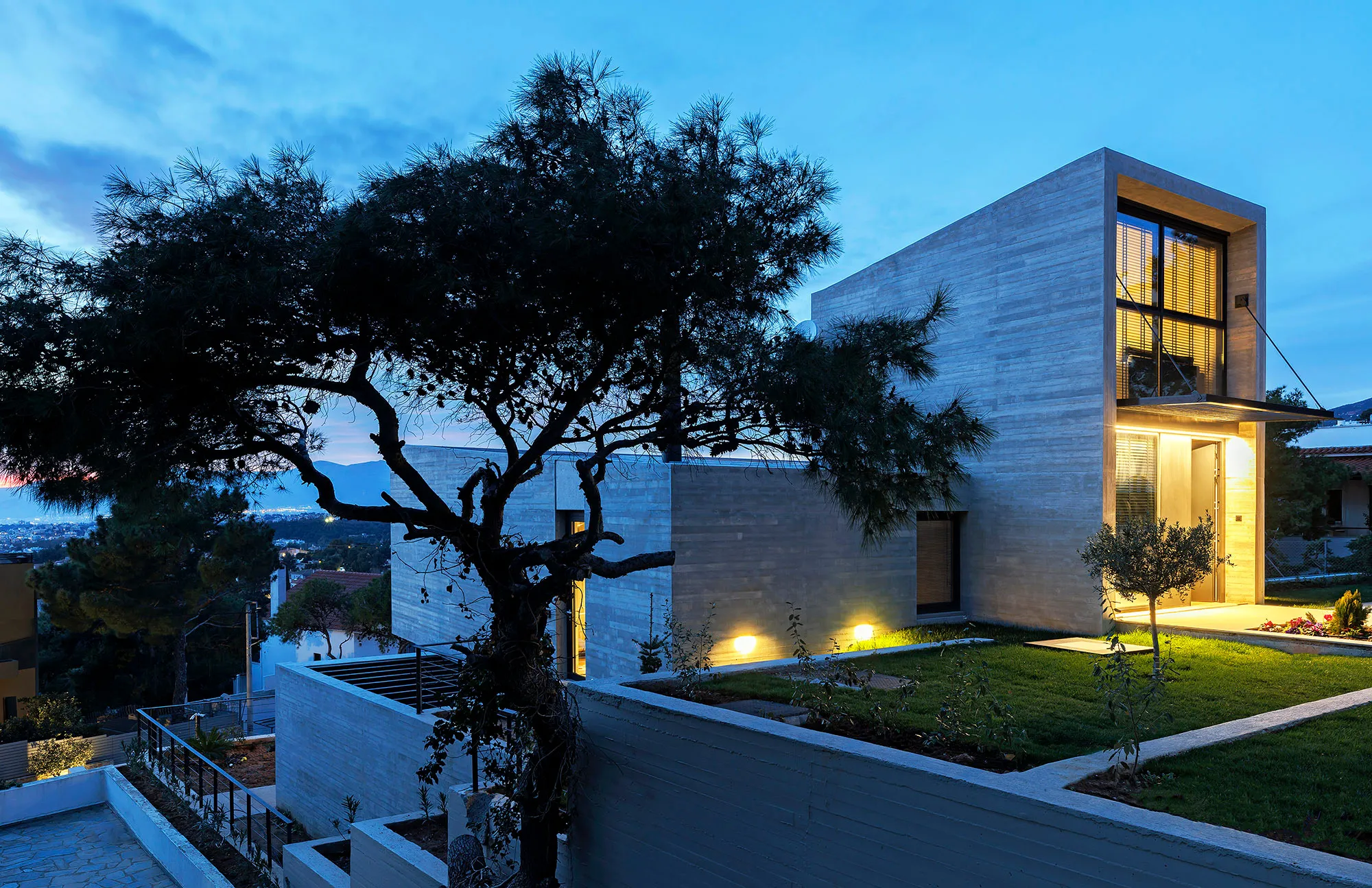 Residence in Penteli: board-marked concrete house at dusk behind a pine tree, with a lit entrance and lawn terraces