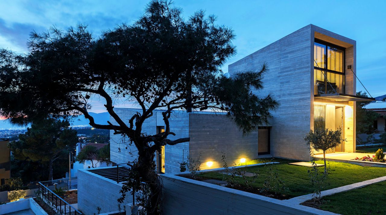 Residence in Penteli: board-marked concrete house at dusk behind a pine tree, with a lit entrance and lawn terraces