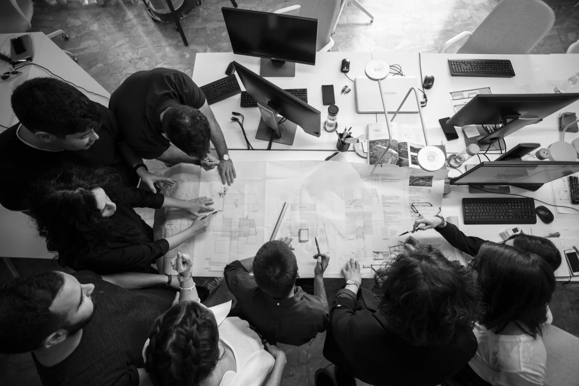 Architects gathered around large printed drawings on a studio table, seen from above in black and white