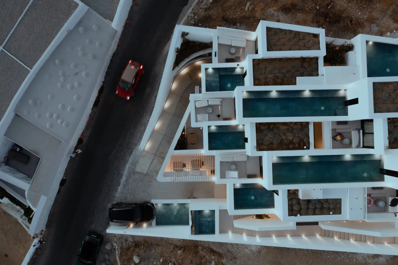 Aerial dusk view of a white boutique hotel on a Tinos slope, with stepped plunge pools and terraces beside a road