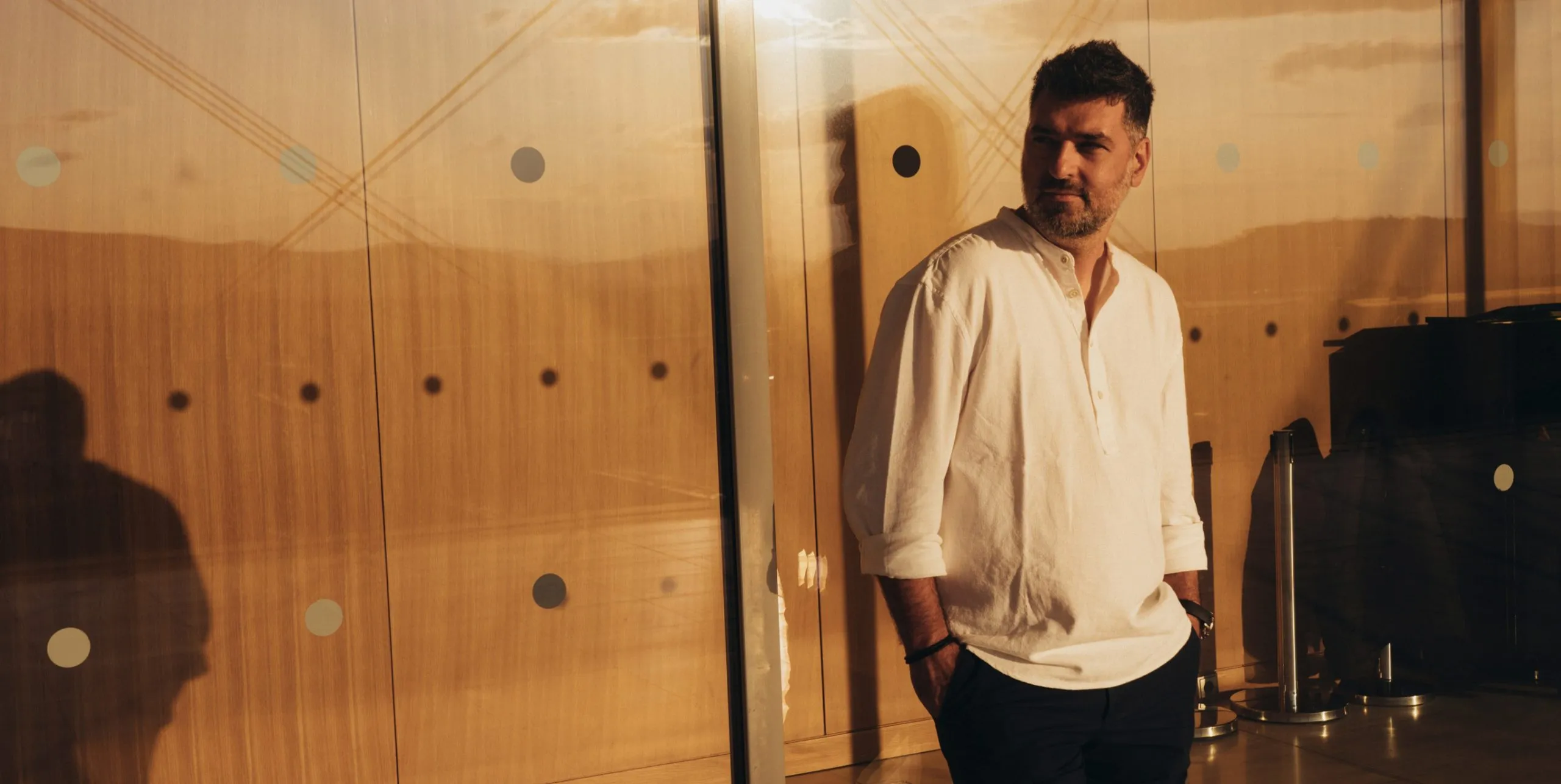 Portrait of Aristides Dallas, founder, standing in a white shirt against a warm timber-panelled wall