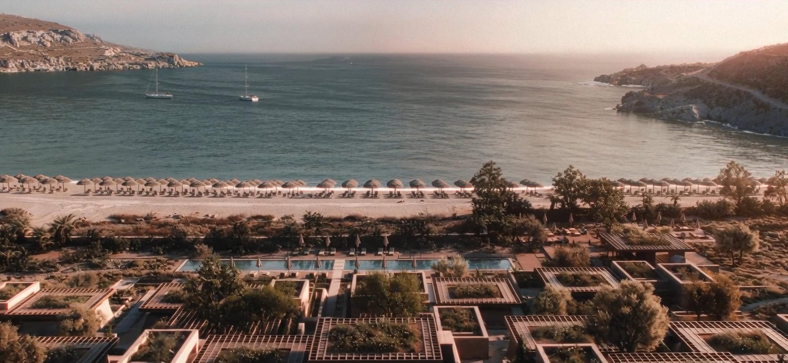 Aerial view of a resort on Andros: planted pergolas and pools behind a sandy beach lined with parasols, facing a calm bay