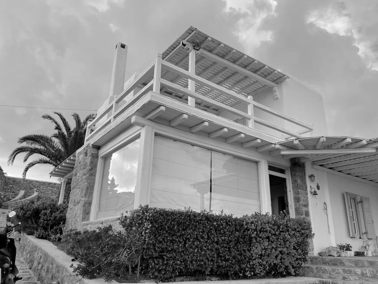 Black-and-white photograph of an existing island house with timber pergolas, a stone base and a palm tree beside it