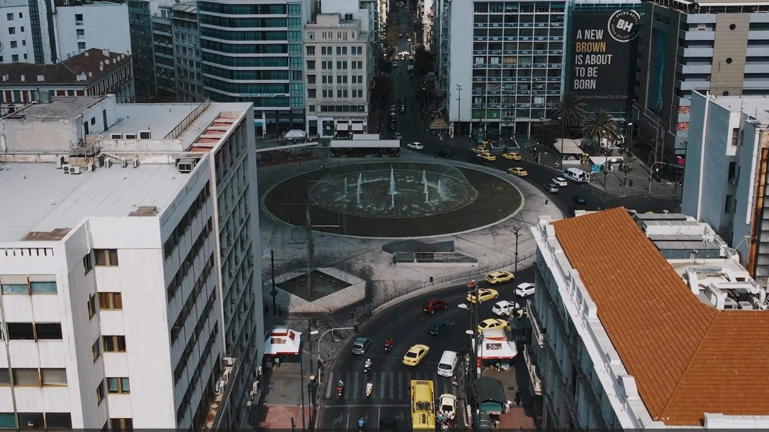 Aerial view of a busy roundabout with a circular fountain, surrounded by dense office and apartment blocks
