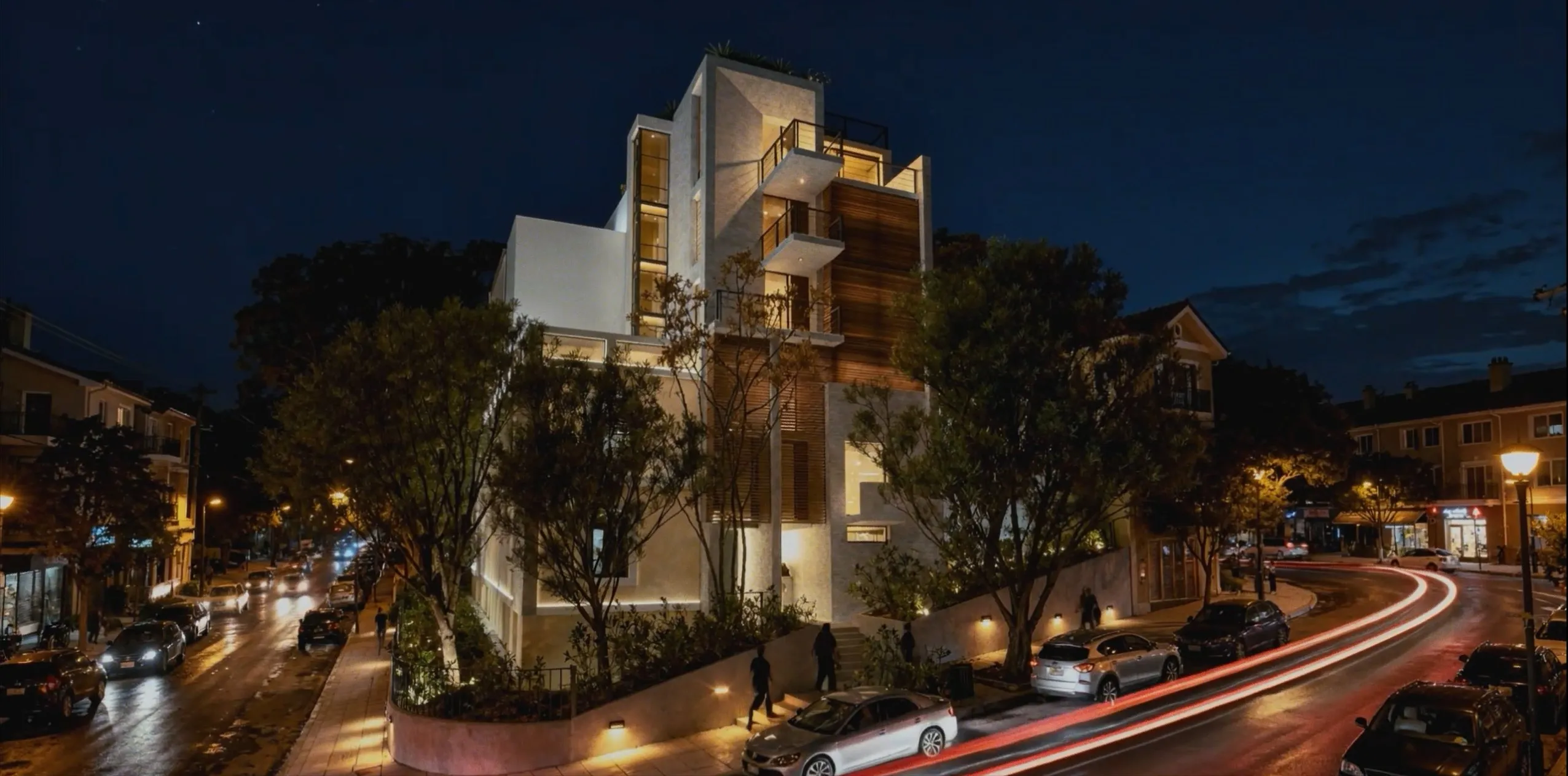 A residential building lit at night above a tree-lined street, with light trails from passing cars