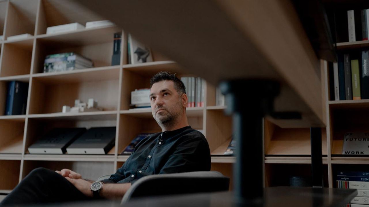 Architect seated in the studio, looking off-camera, framed by wooden shelving full of books and models
