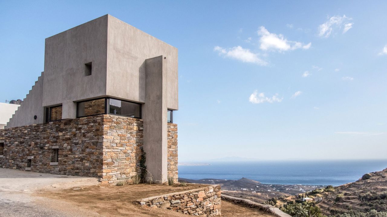 Concrete cubic upper volume on a dry stone base, stepped wall to the left and the sea beyond the Tinos hillside