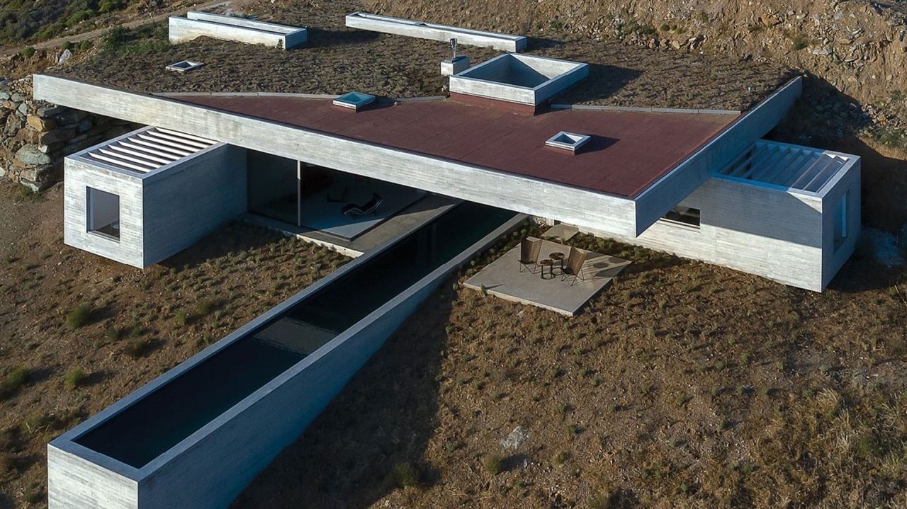 Aerial view of a board-marked concrete house with a red roof plane, rooftop skylights and a long narrow pool cantilevered over the slope