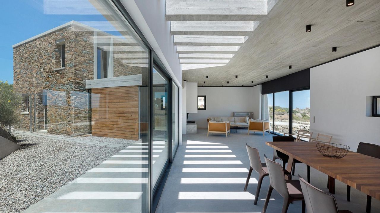 House between the rocks, Volax: living and dining room with a concrete slatted ceiling, glass walls and a stone wing outside