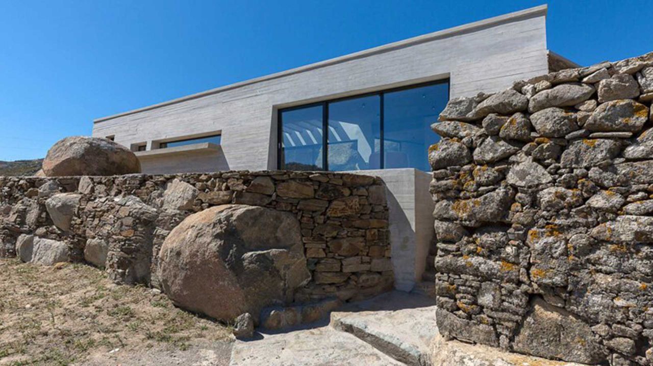House between the rocks, Volax: concrete volume with a large window rising behind dry stone walls and rounded boulders