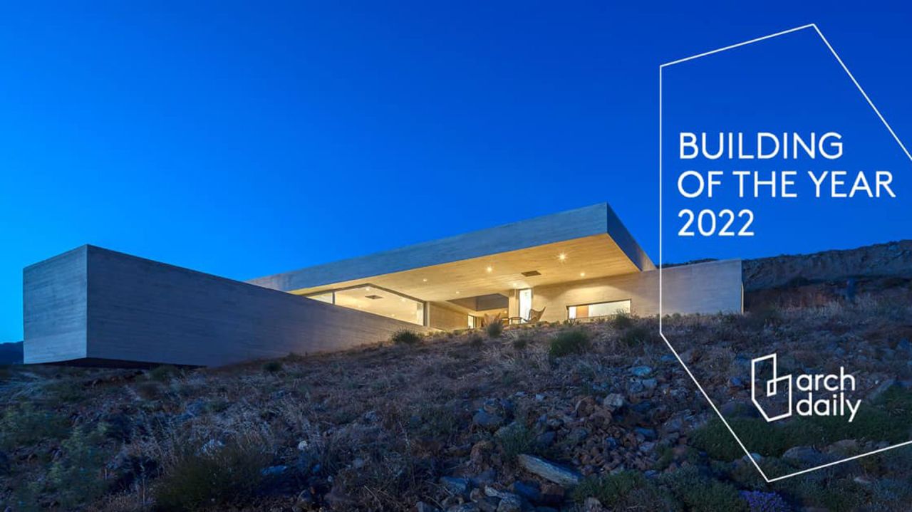 Concrete house with a deep cantilevered roof glowing at dusk on a rocky slope, with the ArchDaily Building of the Year 2022 badge