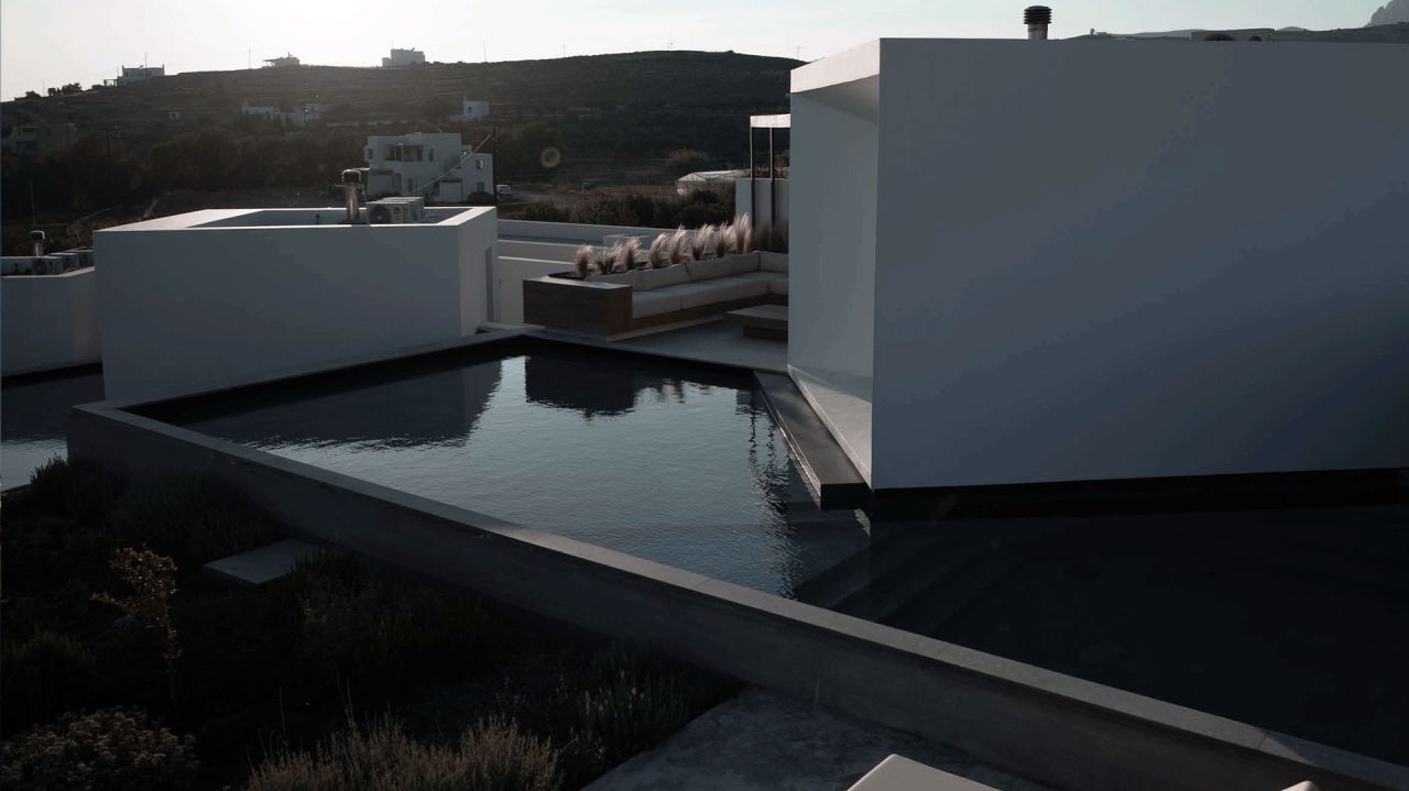 Pnoes Tinos in low evening light: dark pool beside a white cubic volume, rooftop seating with dry grasses and distant village