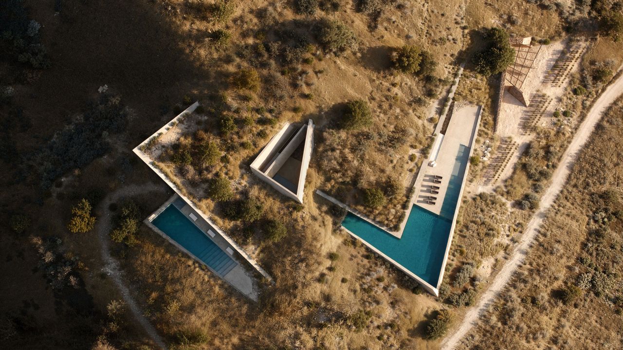 Terra Fracta: aerial view of angular concrete pavilions with two turquoise pools cut into a dry hillside beside a dirt track
