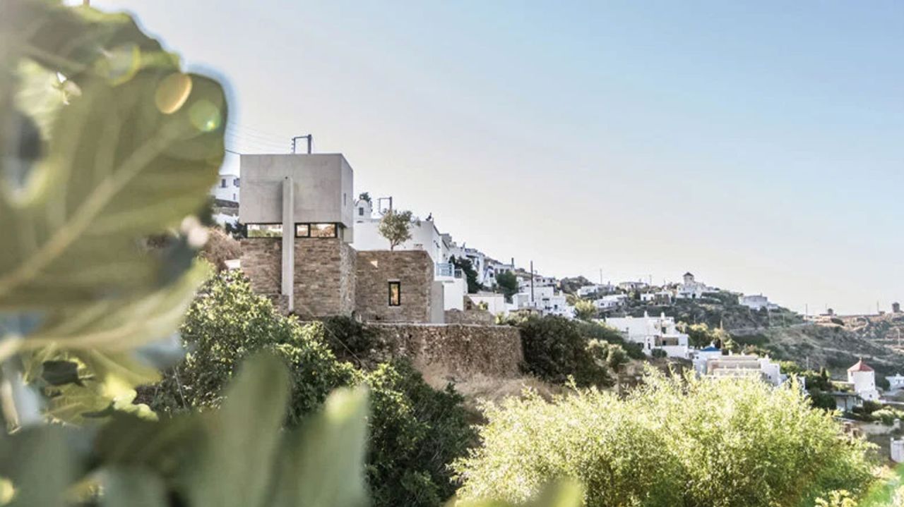 Stone and concrete tower house on a Tinos hillside seen through blurred foliage, with whitewashed village houses beyond