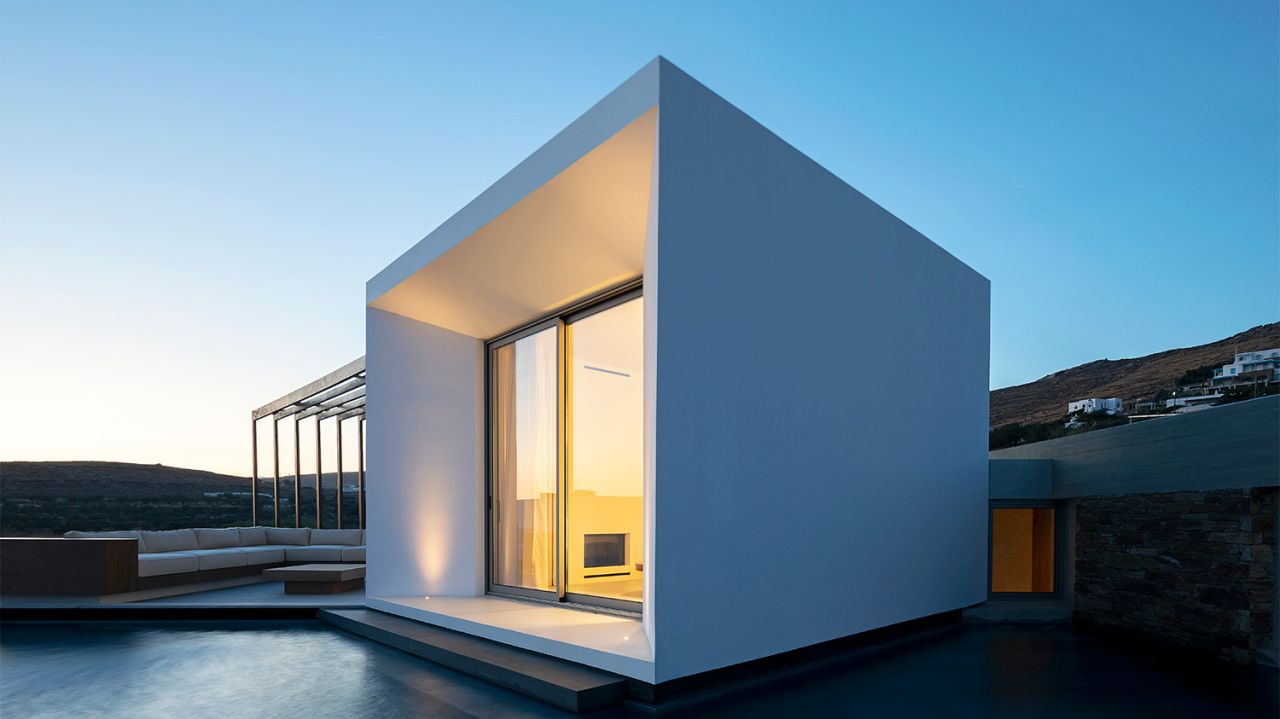 Pnoes Tinos at dusk: white cubic volume with a warmly lit sliding door above a still pool, pergola and lounge seating at left