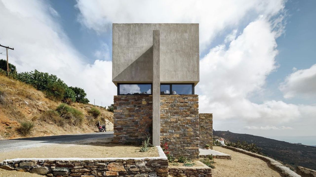 Front view of a concrete cube on a dry stone base with a long ribbon window, beside a hillside road under clouds