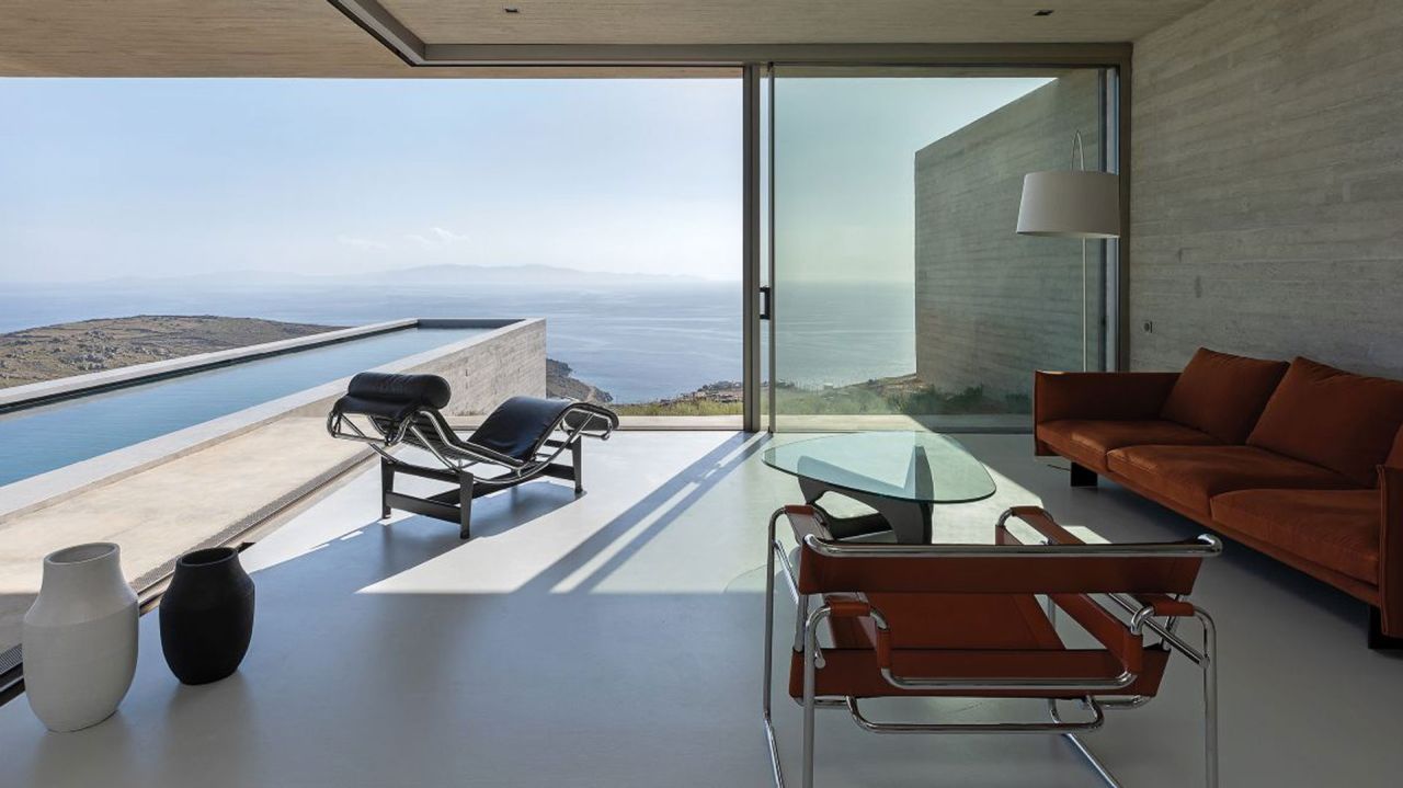 Living room opening to a sea view and long pool, with a chrome lounge chair, leather armchair, rust-coloured sofa and glass table