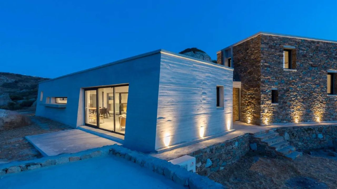 Board-marked concrete wing and a dry stone wing of a house lit by uplights at night, with a terrace and stone steps