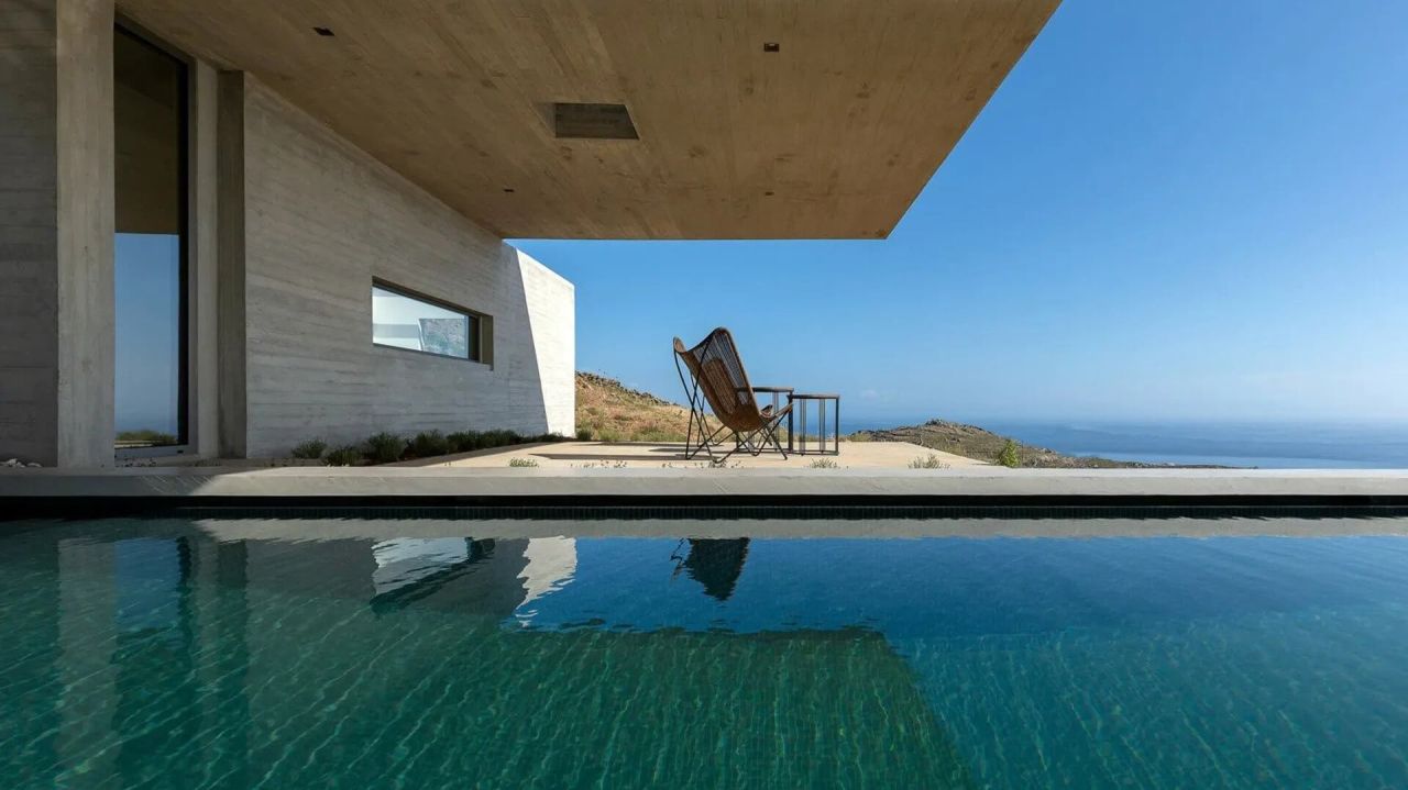 View across a green pool to a concrete cantilevered roof, a butterfly chair on the terrace and the sea on the horizon