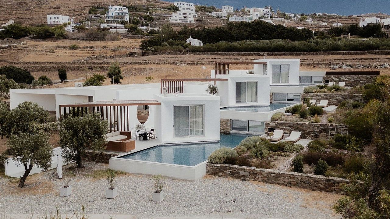 Pnoes Tinos: cluster of white cubic houses with pools, pergolas and stone terraces in front of a Tinos village and olive trees