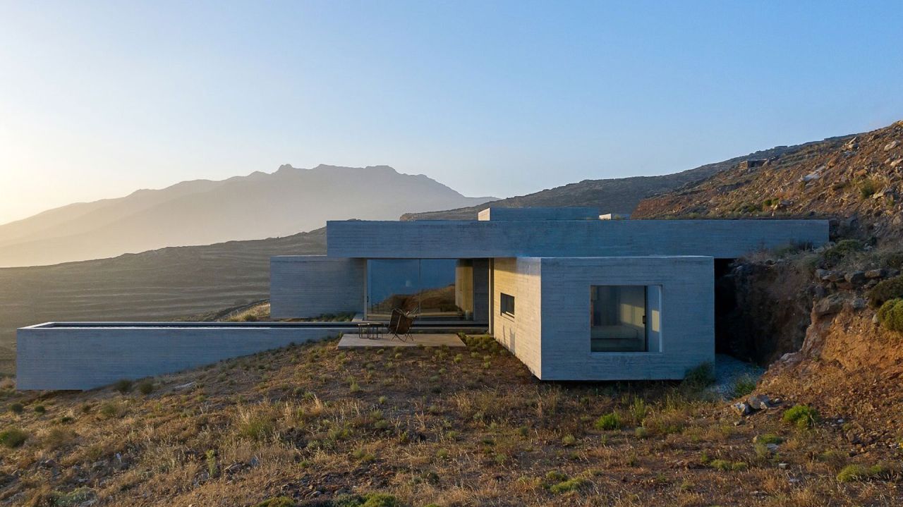 Board-marked concrete house stepping down a mountain slope at sunset, a long pool extending to the left and distant mountains