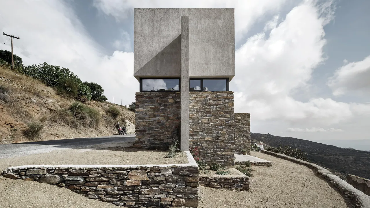 A Touch of New in Triantaros, Tinos: concrete volume on a stone base with a tall concrete pillar, beside a hillside road