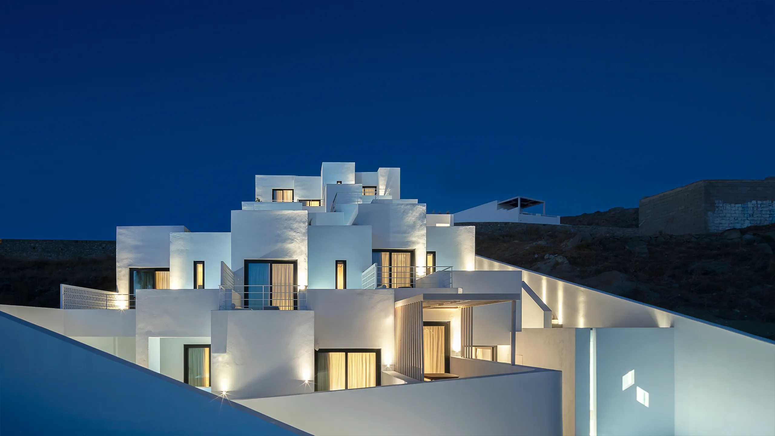 Cubic Hotel, Mykonos: stepped white cubic volumes glowing with warm light against a deep blue night sky
