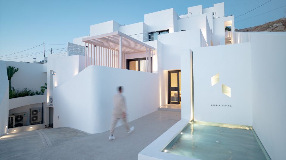 Cubic Hotel entrance at dusk, with a walking figure, a pergola, the CUBIC HOTEL sign and a shallow reflecting pool