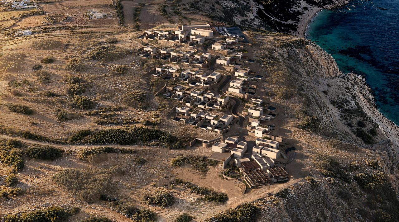 Aerial view of Erema Milos: clusters of low, earth-toned volumes stepping down a dry hillside above the sea at Cape Chalaka