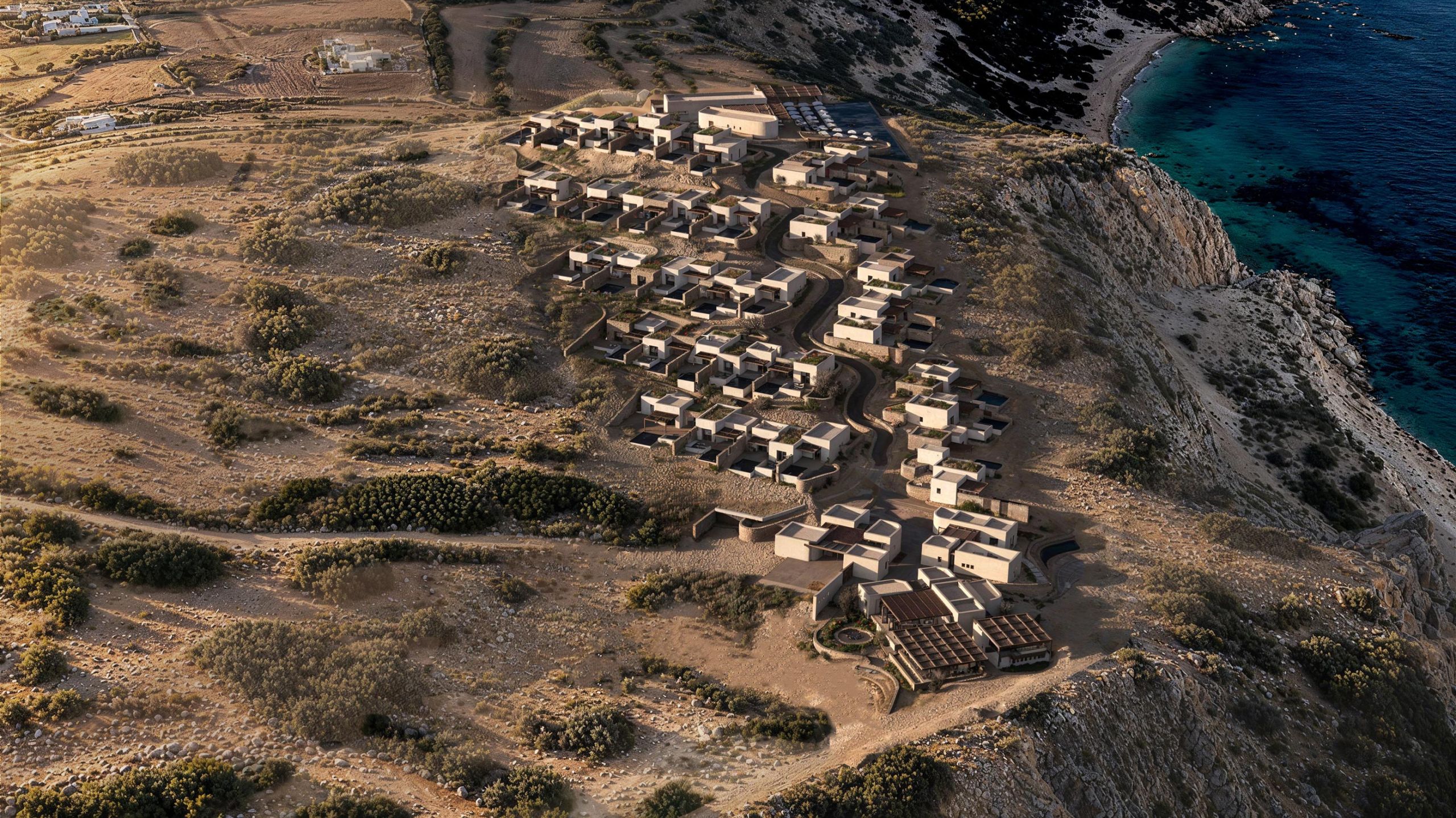 Aerial view of Erema Milos: clusters of low, earth-toned volumes stepping down a dry hillside above the sea at Cape Chalaka