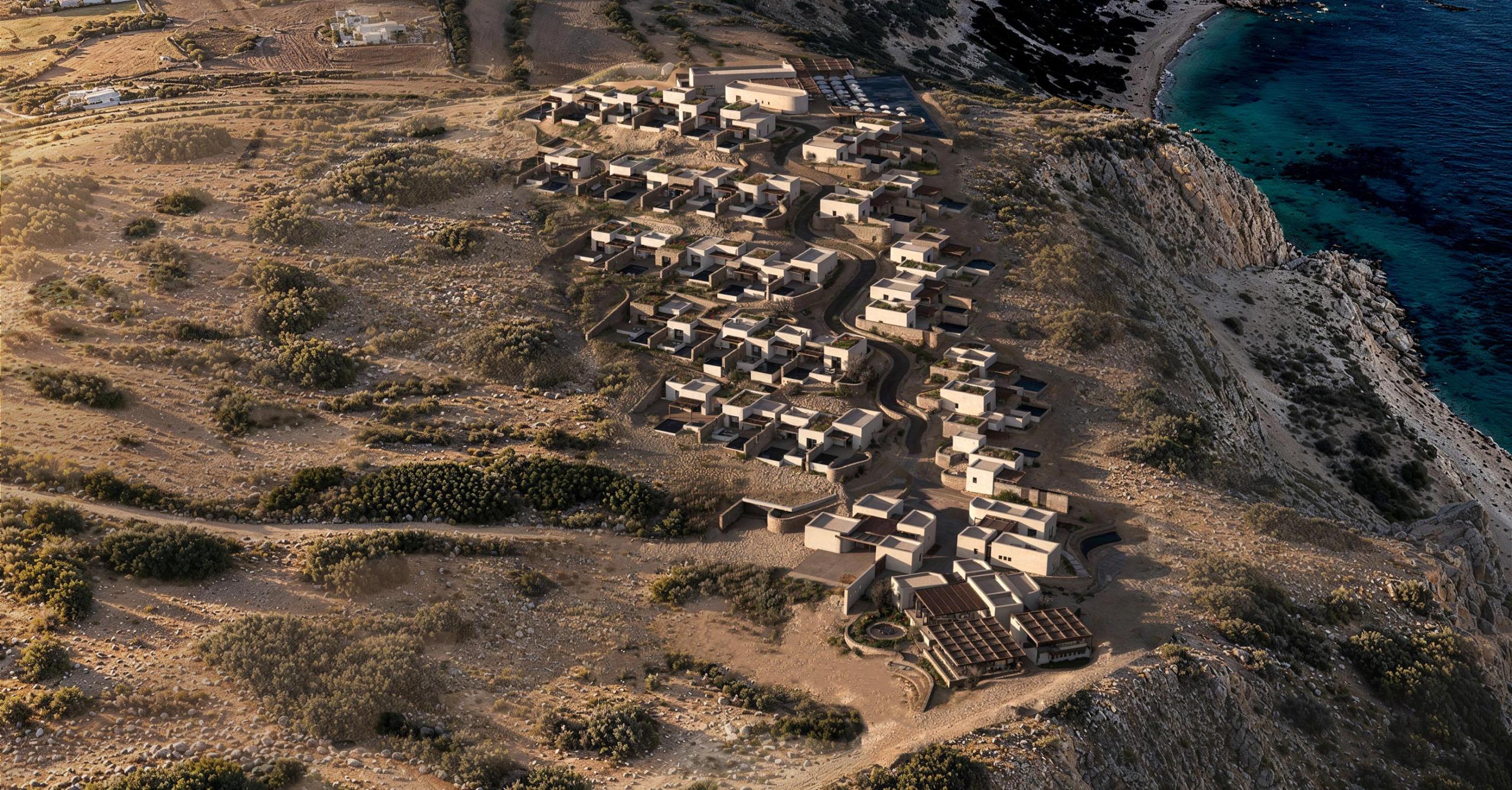Aerial view of Erema Milos: clusters of low, earth-toned volumes stepping down a dry hillside above the sea at Cape Chalaka