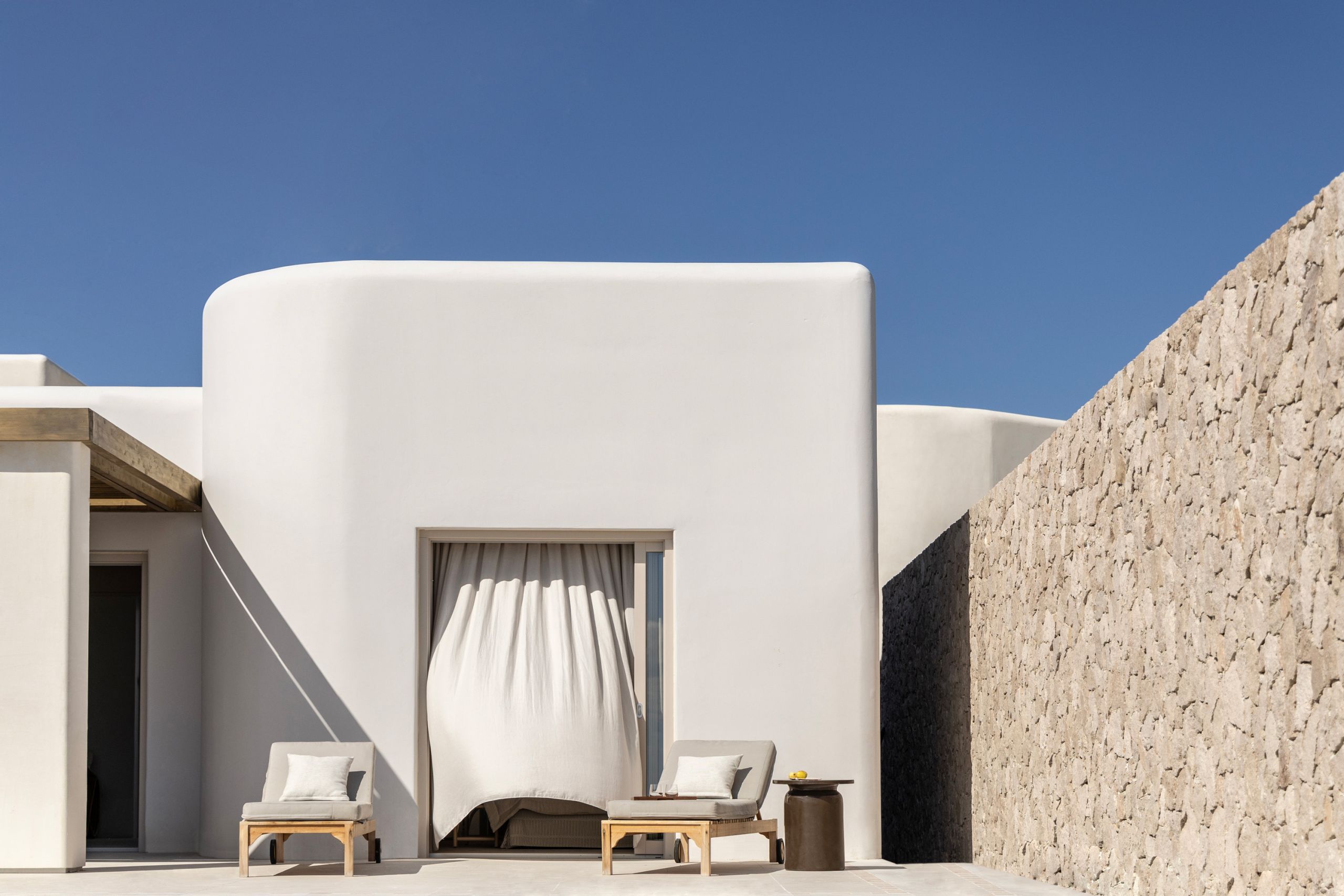 A white plastered suite with a curtained doorway and two loungers, beside a dry-stone wall