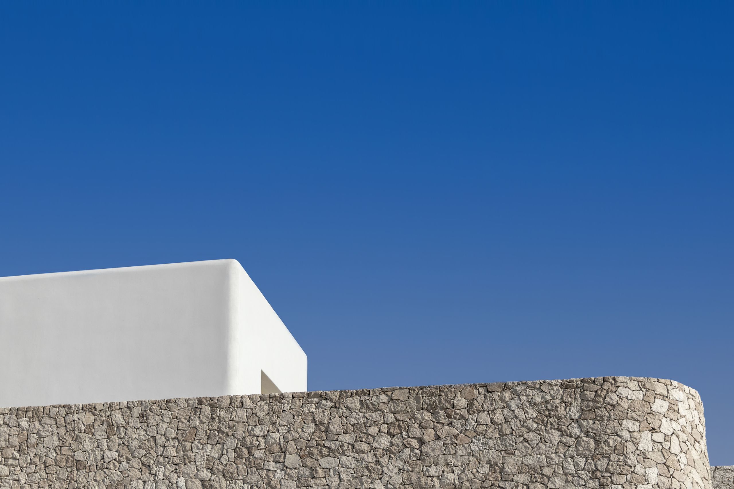 A white volume above a curved dry-stone wall against a clear blue sky
