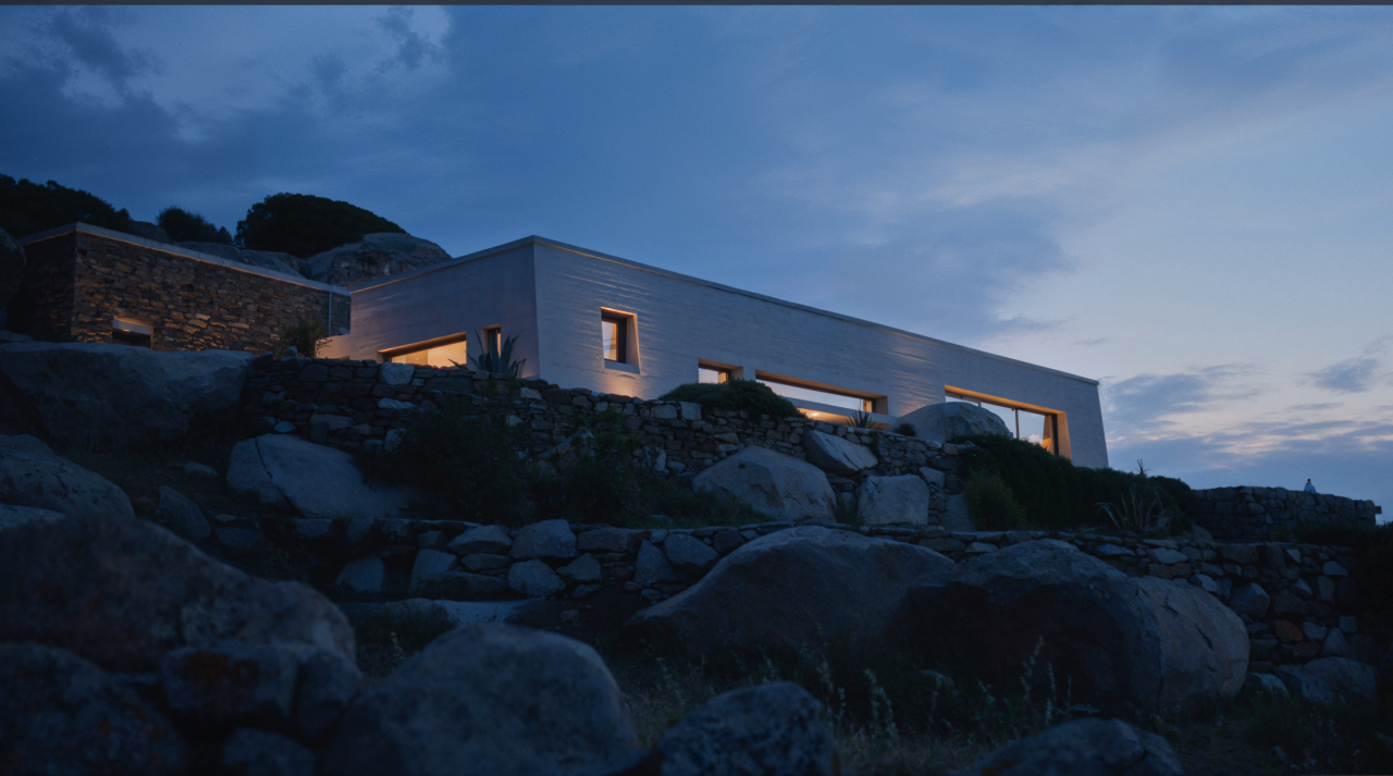 House in Volax at dusk: long pale volume with warm lit windows set above dry-stone walls and boulders under a cloudy sky