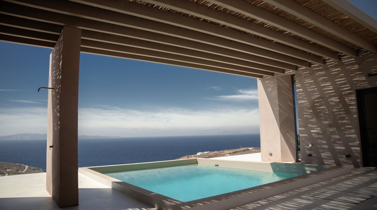 A swimming pool under a timber pergola, open to the Aegean horizon