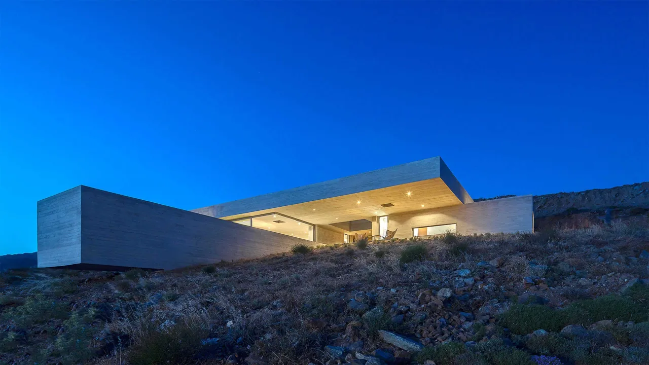 The Lap Pool House featured view: illuminated concrete house with a deep cantilevered roof on a Tinos hillside under a deep blue sky