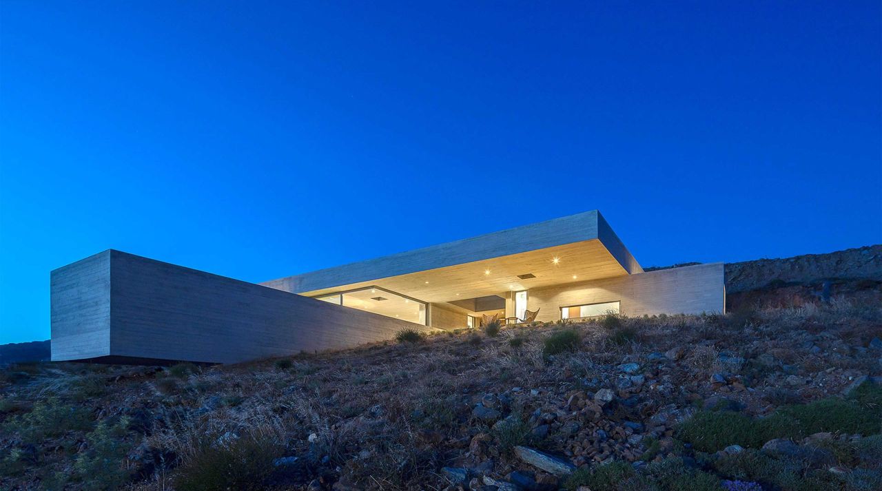 The Lap Pool House featured view: illuminated concrete house with a deep cantilevered roof on a Tinos hillside under a deep blue sky