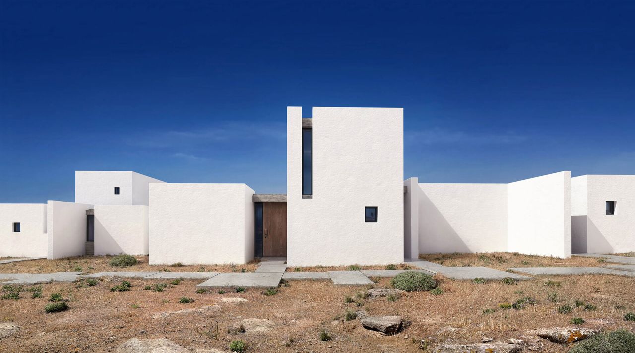 Out of the Box, Mykonos: row of white cubic residences with a timber front door and stepping-stone paving across the dry hillside under a deep blue sky