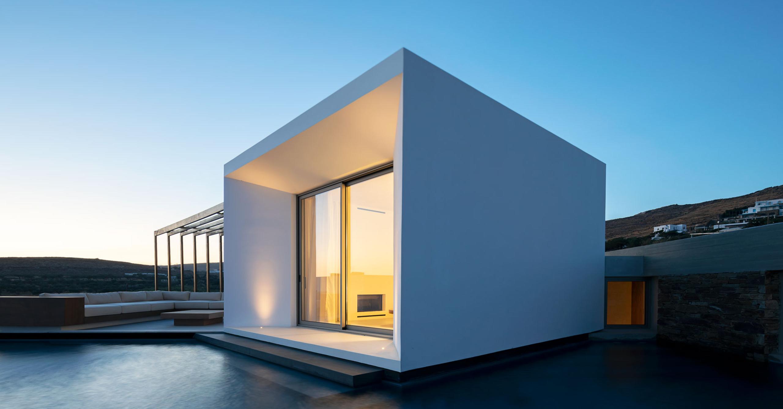Pnoes Tinos: white cubic villa with a deep overhang and glowing glazing above a dark pool at dusk, pergola and lounge to the left