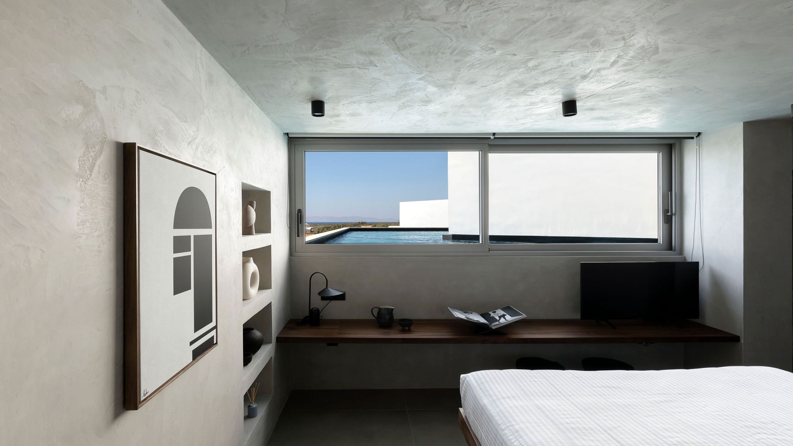 Pnoes Tinos bedroom: plaster-finish interior with a bed, wall niches, framed print and a person leaning at a timber desk by the window