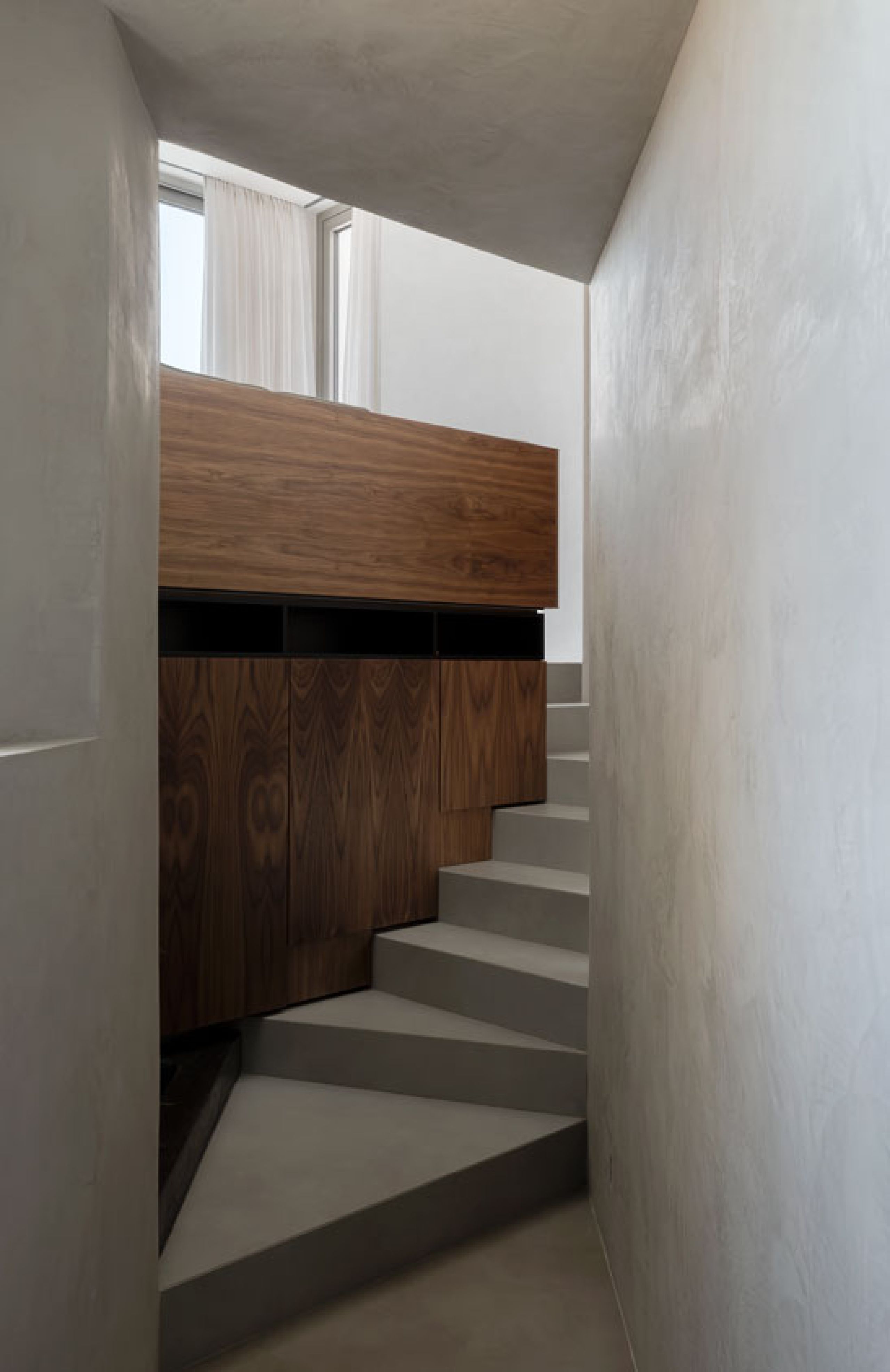 Pnoes Tinos interior stair of grey microcement steps beside walnut cabinetry, with a bright curtained window above