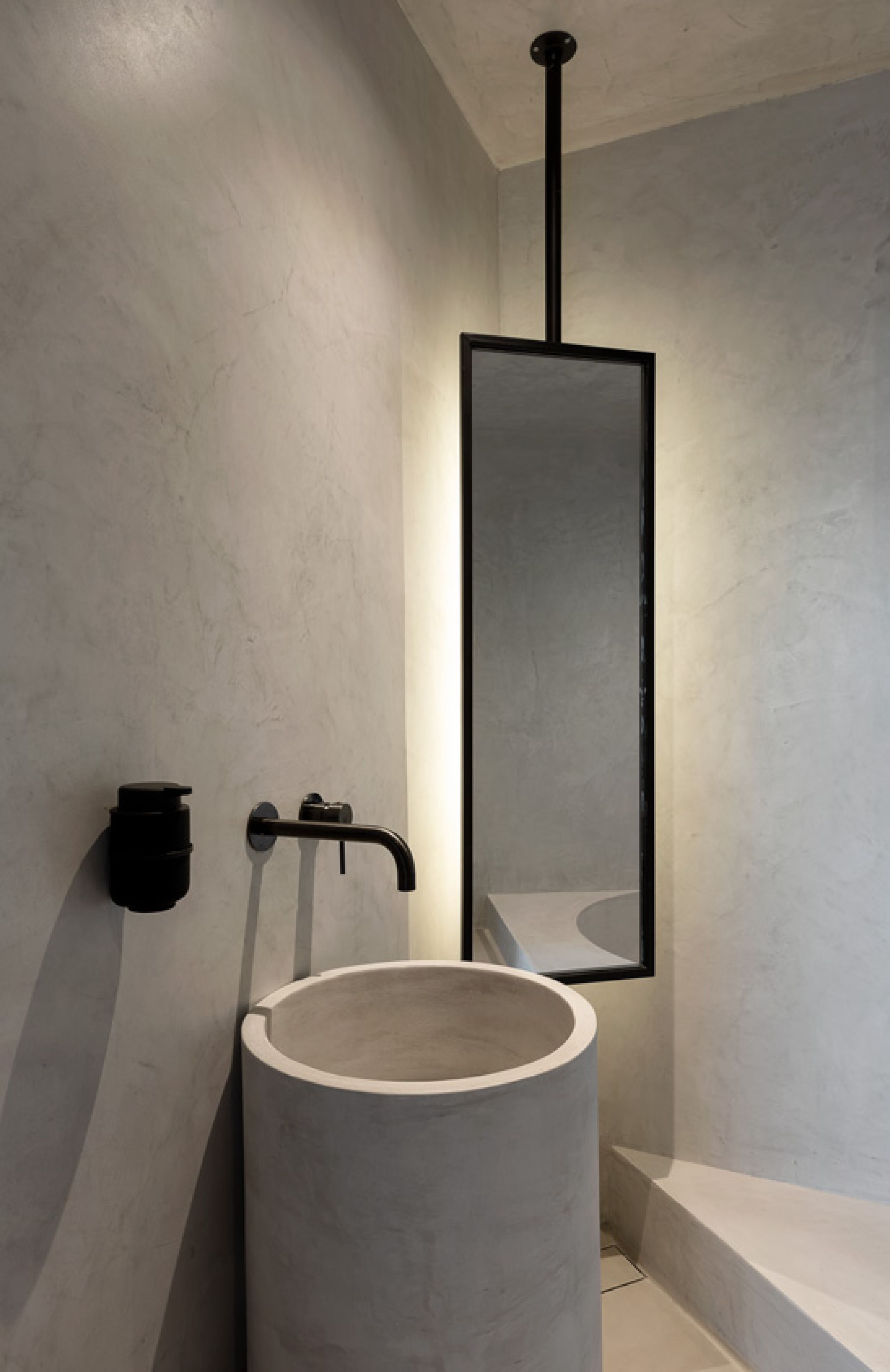Pnoes Tinos bathroom corner: round concrete-finish basin, black wall tap and a ceiling-hung framed mirror with backlight