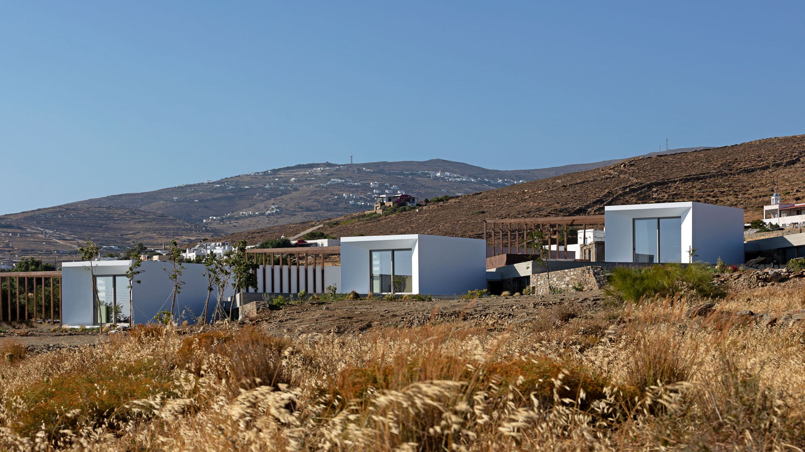 Pnoes Tinos: cluster of white cubic villas with glazed fronts and timber pergolas on the dry hillside, with Tinos hills behind
