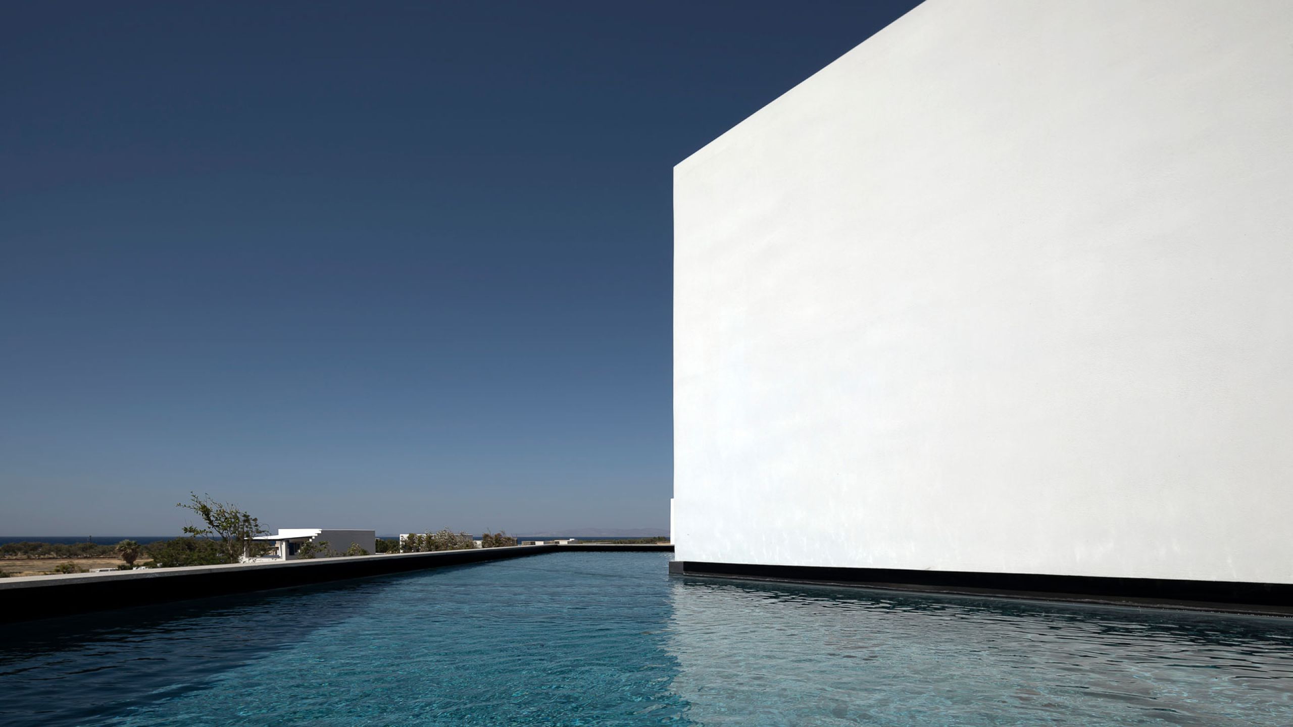 Pnoes Tinos: long narrow pool reflecting a tall white wall, with low villas and the sea on the horizon under a deep blue sky