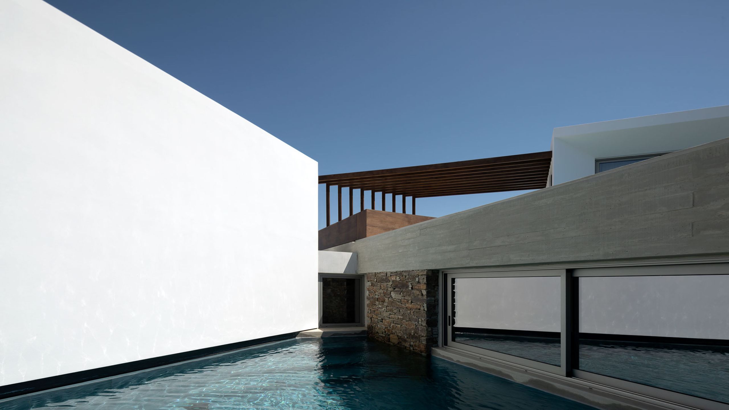 Pnoes Tinos: pool running between a white wall and a board-marked concrete wall with a stone base, with a timber pergola above