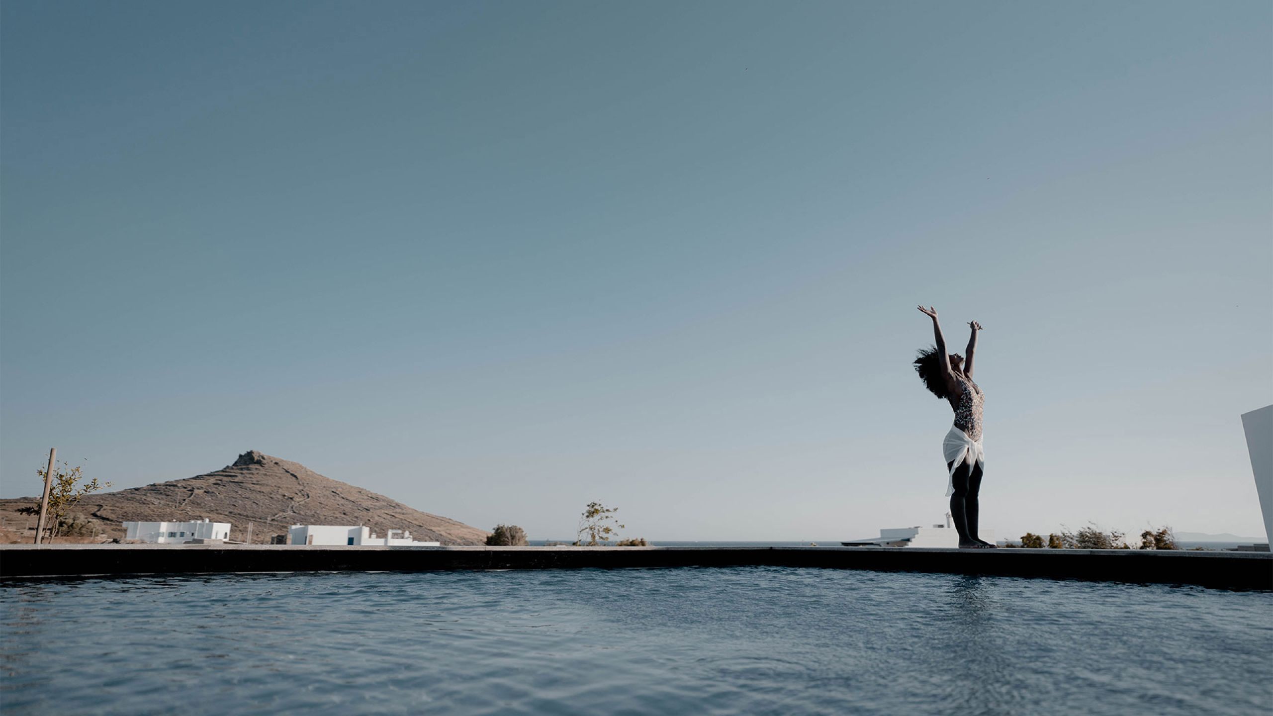 Person with raised arms standing at the dark edge of a Pnoes Tinos pool, with white buildings and a hill in the distance