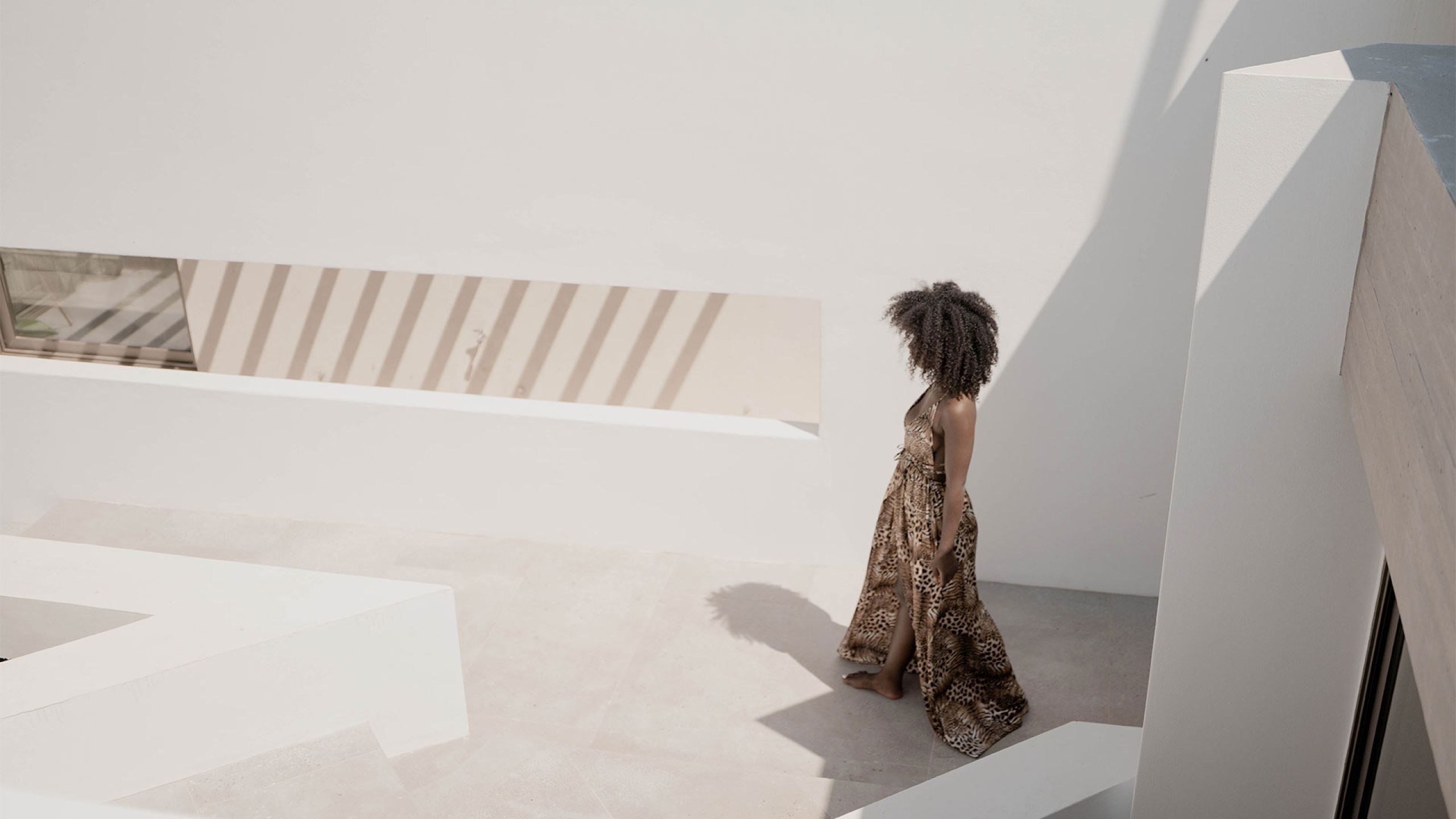 Pnoes Tinos courtyard: a person in a patterned dress walks across pale paving among white walls casting striped shadows